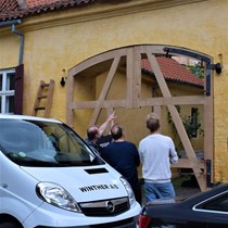 En tømrersvend og en snedkersvend har med hjælp fra smeden klodset de to portrammer op i porthullet og de nye håndsmedet hængsler er hængt på de oprindelige stabler.